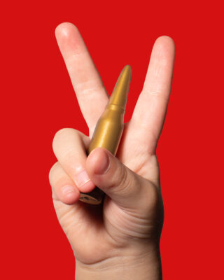 Photograph with an intense red background and a child's hand making the peace sign with a large bullet held in its hand.