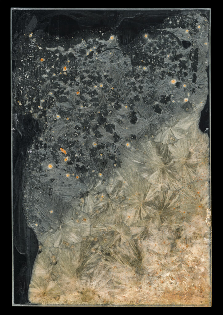 Photograph with a black background of crystalized feathery formations in a light grey turning to a peachy-red at the bottom right.