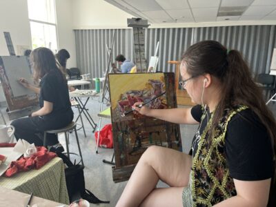 Photograph of a studio with people seated an easels painting a still life.