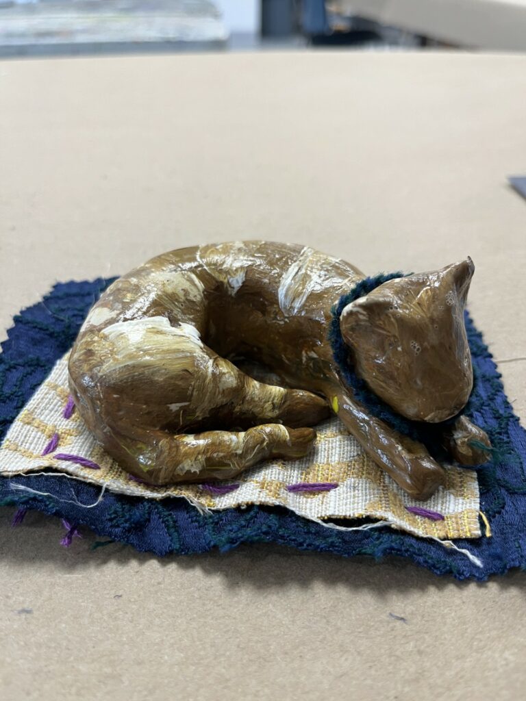 Photograph of a brown ceramic cat, curled up sleeping on a blue and cream rug.