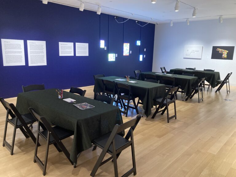 Photograph of a gallery with tan floors, a dark blue wall on the left and a white wall on the right with artwork. In the center are long black covered tables with black chairs and art supplies on top.