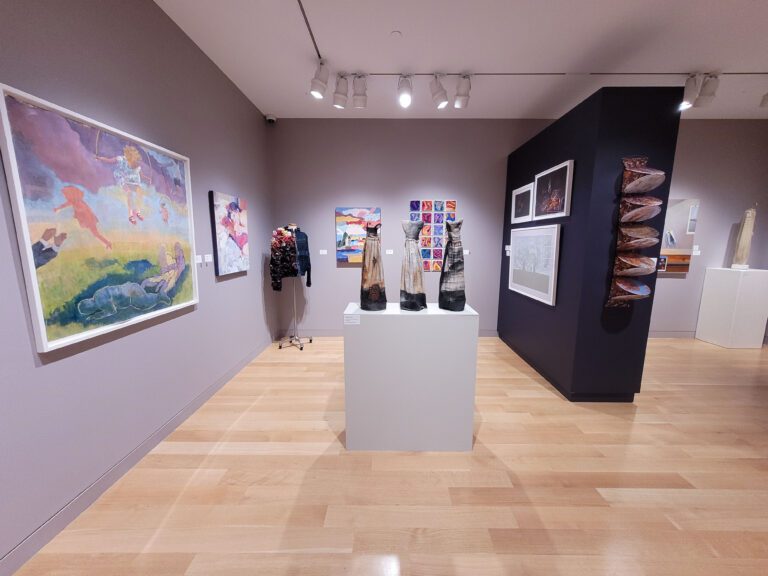 Gallery installation shot with a tan floor, mauve walls, and sections of deep purple walls. There are various artworks hanging from the walls.