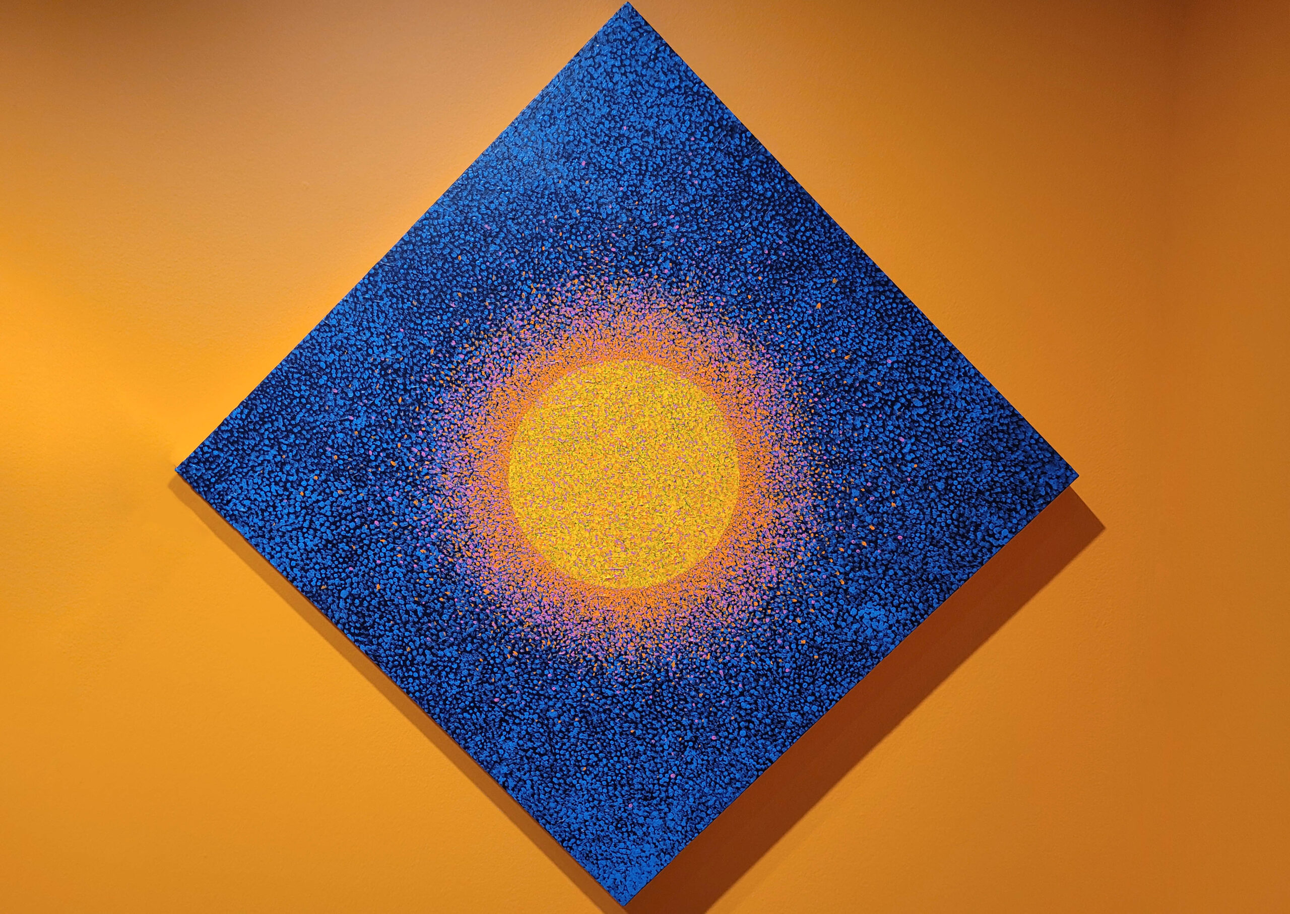 Diamond painting with a black and blue speckled background and a bright yellow orb in the center, surrounded by a speckled orange radiating band.