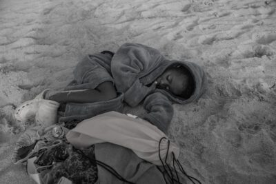 Black and white photograph of a child curled up sleeping in the sand.