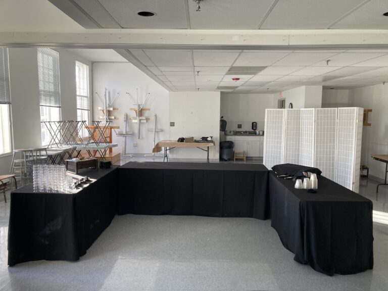 Photograph of a room with white walls and windows to the left. In the center in a 'U' shape are three black tables with supplies for food service.