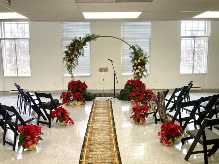 Photograph of an event space with white walls and large windows, a rug down the center leading to an arch, and several rows of black chairs on either side of the rug with poinsettia flowers.