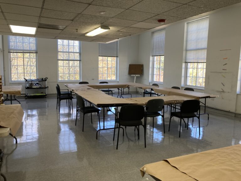 Photograph of a studio room with windows and a square of brown covered tables in the center and chairs around it.