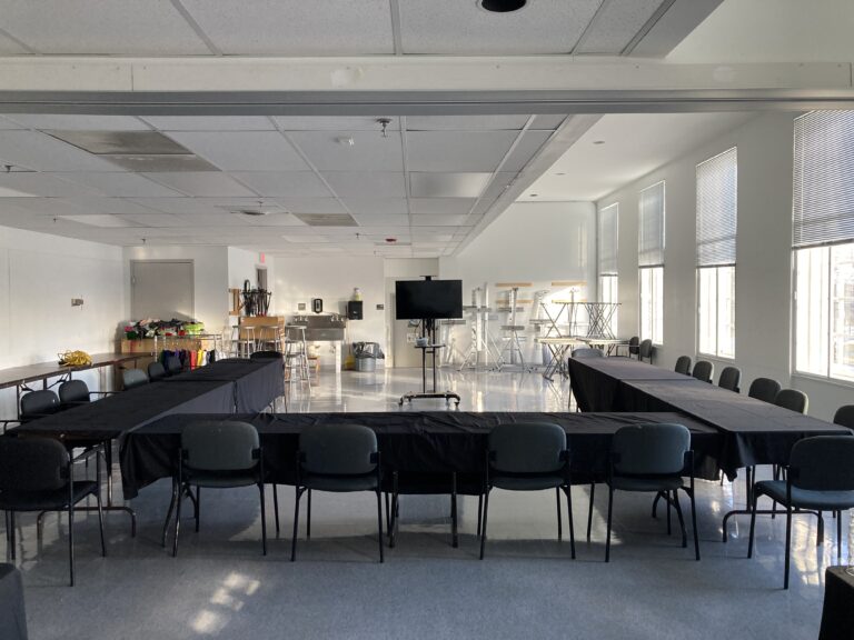 Photograph of a room with white walls and windows to the right. In the center in a 'U' shape are six black tables with chairs all facing a tv in the back center.