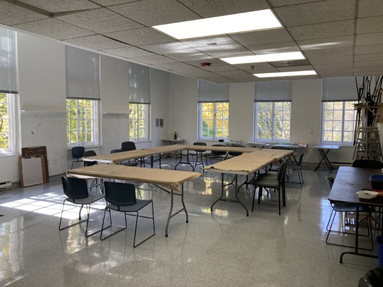 Photograph of a studio room with windows and a square of brown covered tables in the center and chairs around it.