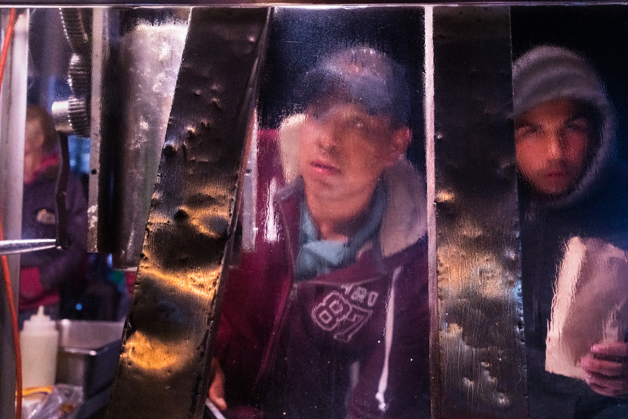 Photograph of a metalic, glowing surface with reflections of two men. The one in the center has a red sweater and baseball hat while the man on the right has a dark hoodie and holding a paper bag.