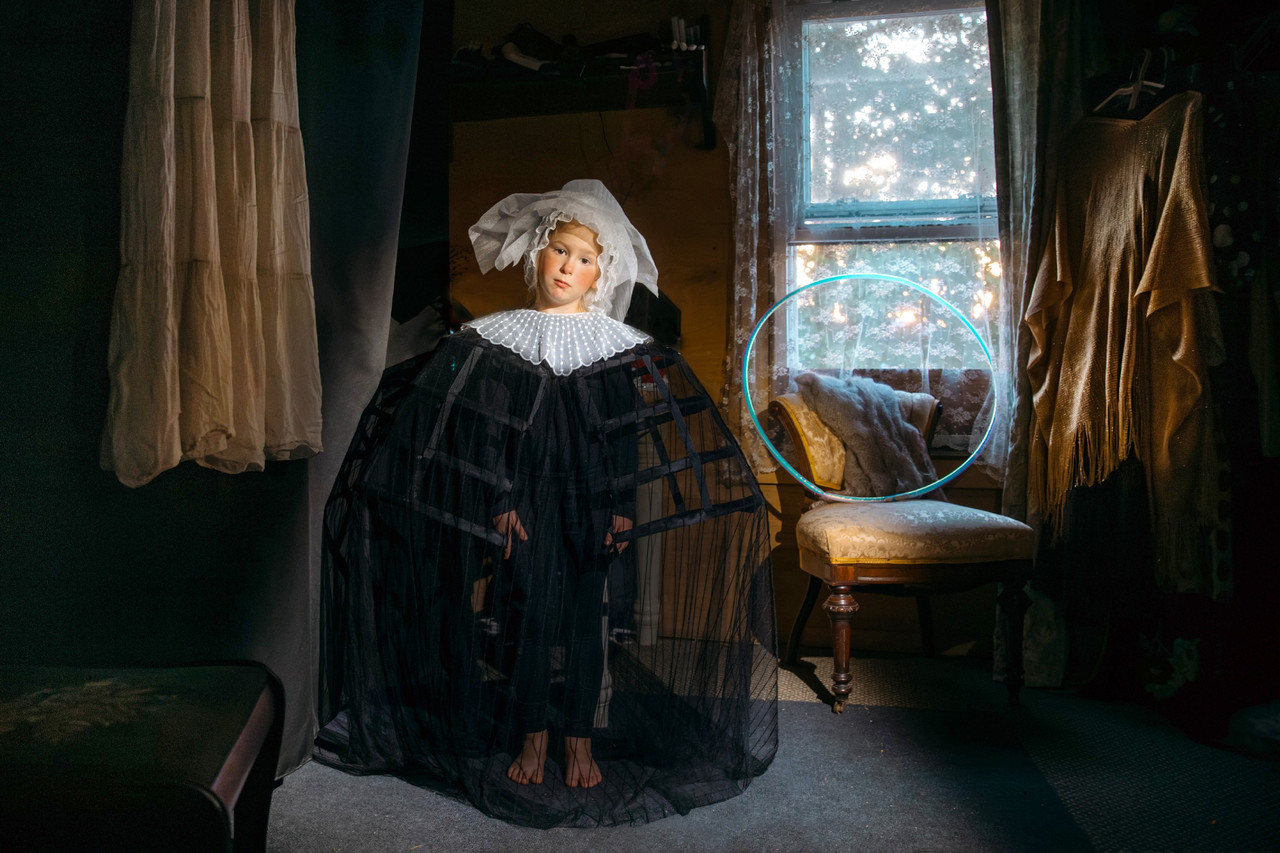 Photograph of an interior room with fabric hangings and a window to the center right with a chair and teal hula-hoop under it. There is a child figure to the left of the window and chair in a wide black see through outfit, a wide bonnet, and an unemotional expression on their face.